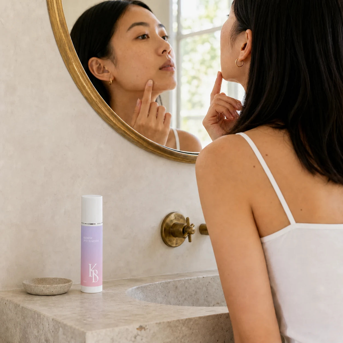 Woman dotting Kaloneo Beauty Blemish Spot Remover onto her jawline at a stone vanity beneath a round brass mirror, the 30ml bottle in the foreground