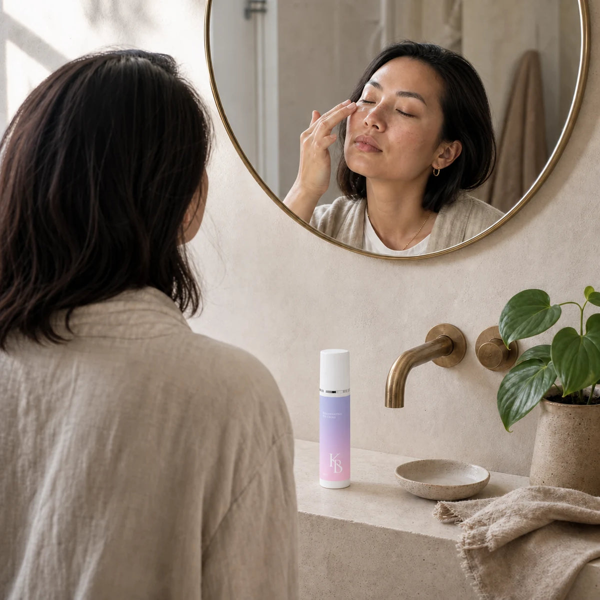 Woman patting Kaloneo Beauty Rejuvenating Eye Cream along her under-eye at a stone vanity beneath a round brass mirror, the 30ml bottle in the foreground