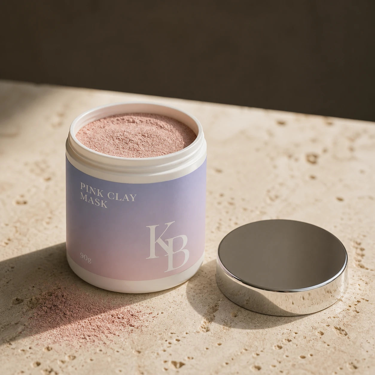 The open Kaloneo Beauty Pink Clay Mask jar showing the fine dry dusty-rose clay powder inside, its chrome lid resting on the travertine beside it and a little powder tapped out onto the stone