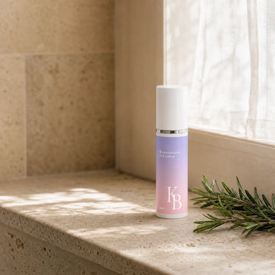 Kaloneo Beauty Rejuvenating Eye Cream 30ml standing on a travertine window ledge in morning light, with a sprig of rosemary beside it and a sheer curtain behind