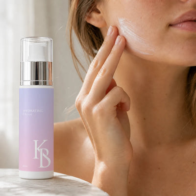 Woman smoothing Kaloneo Beauty Hydrating Cream along her jawline in soft window light, the 60ml bottle in the foreground