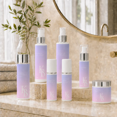 Kaloneo Beauty Hydrating Cream 60ml standing with the complete seven-product Kaloneo Beauty range on a travertine bathroom vanity in morning light, an olive branch in a glass vase, folded towels and a round brass mirror behind