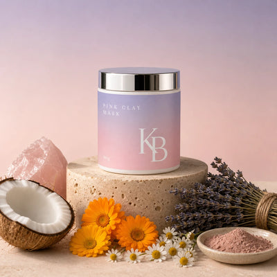 Kaloneo Beauty Pink Clay Mask 90g on a travertine plinth with rose quartz, fresh coconut, calendula and chamomile flowers, dried lavender and a dish of raw pink clay powder, the botanicals used in the formula