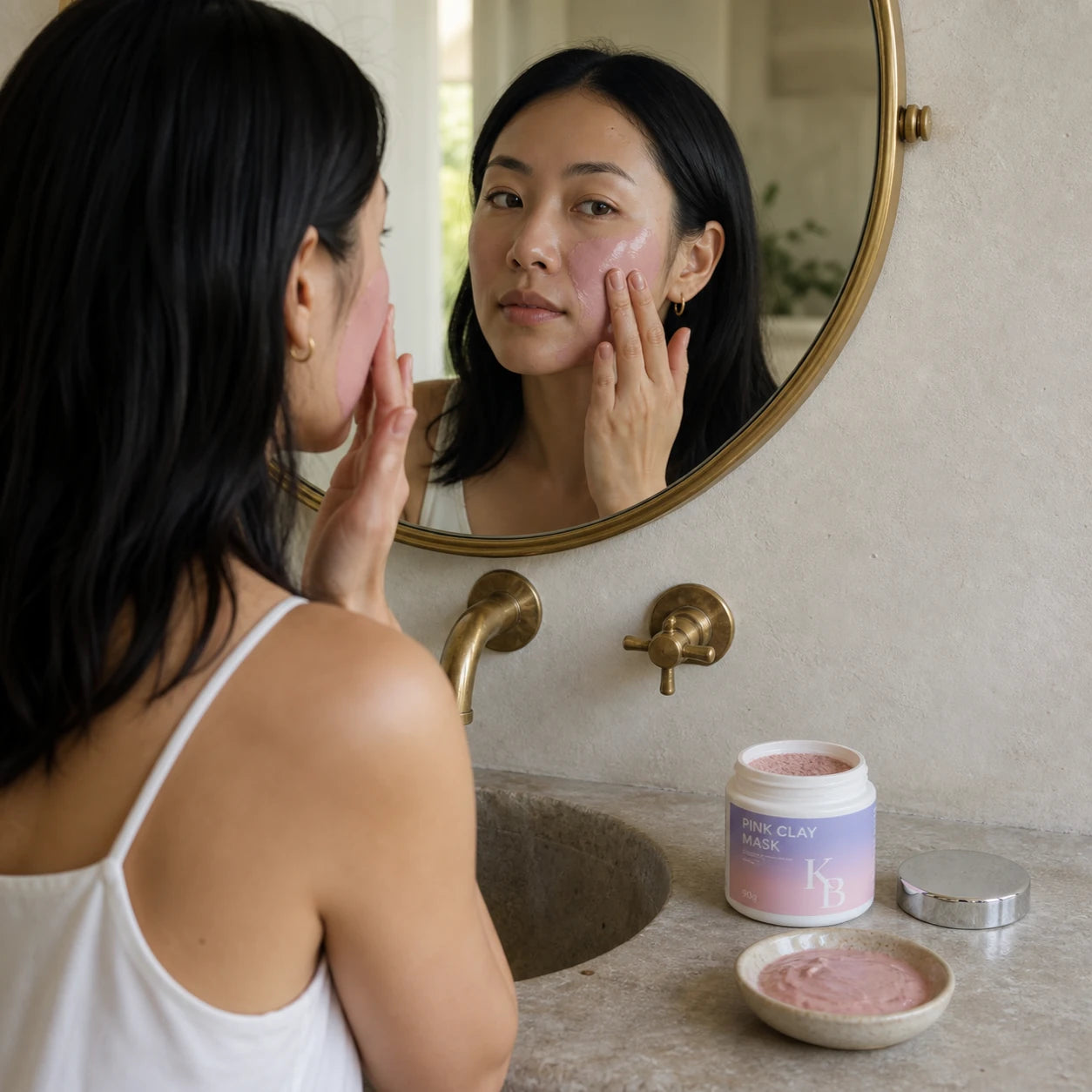 Woman smoothing freshly mixed Kaloneo Beauty Pink Clay Mask across her cheek at a stone basin beneath a round brass mirror, the open jar and a dish of mixed clay paste on the vanity beside her